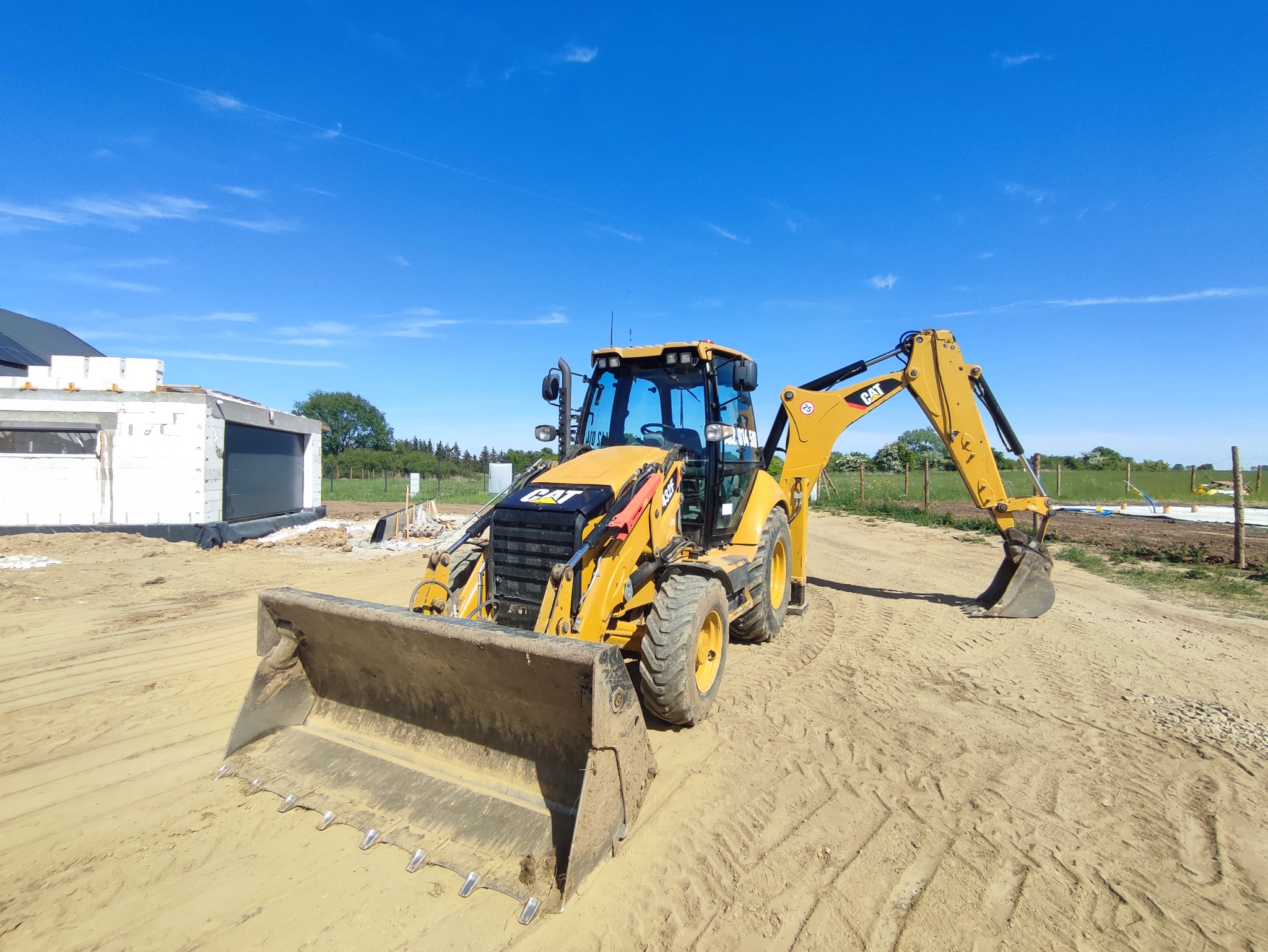 Żółta koparko-ładowarka CAT 432F2 na placu budowy z widocznym budynkiem w stanie surowym otwartym i świeżo wyrównanym terenem.