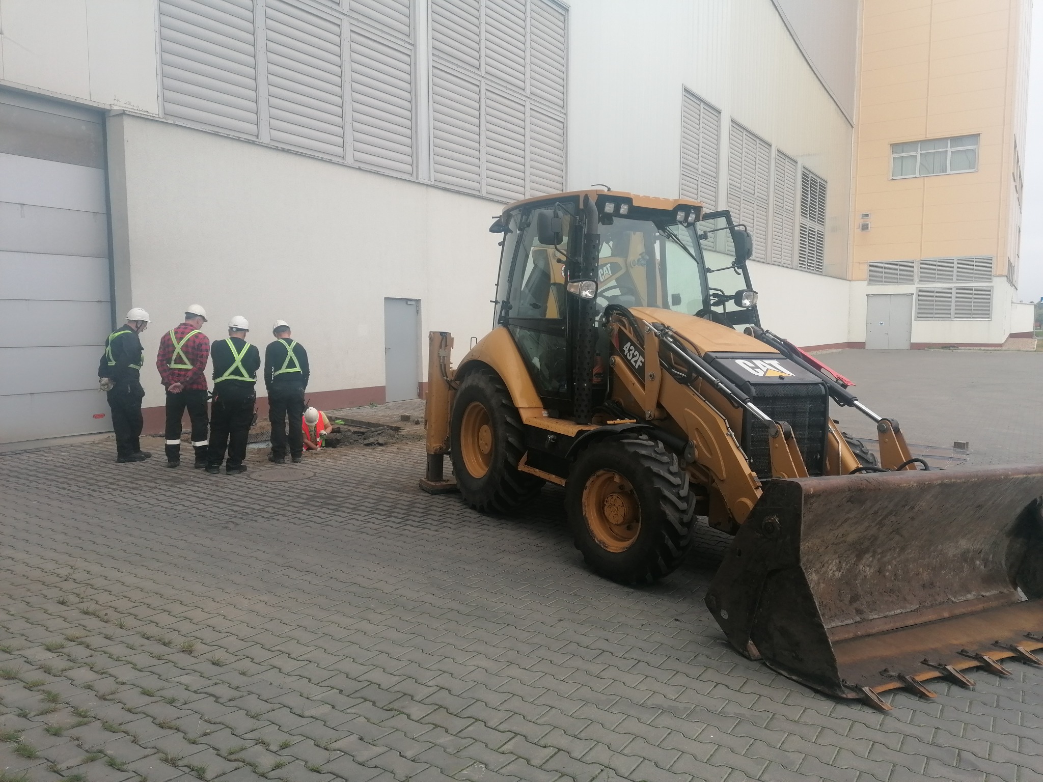 Żółta koparko-ładowarka CAT 432F zaparkowana na kostce brukowej obok grupy robotników w kaskach, jeden z nich w kamizelce odblaskowej pracuje w wykopie, na tle budynku przemysłowego z licznymi...