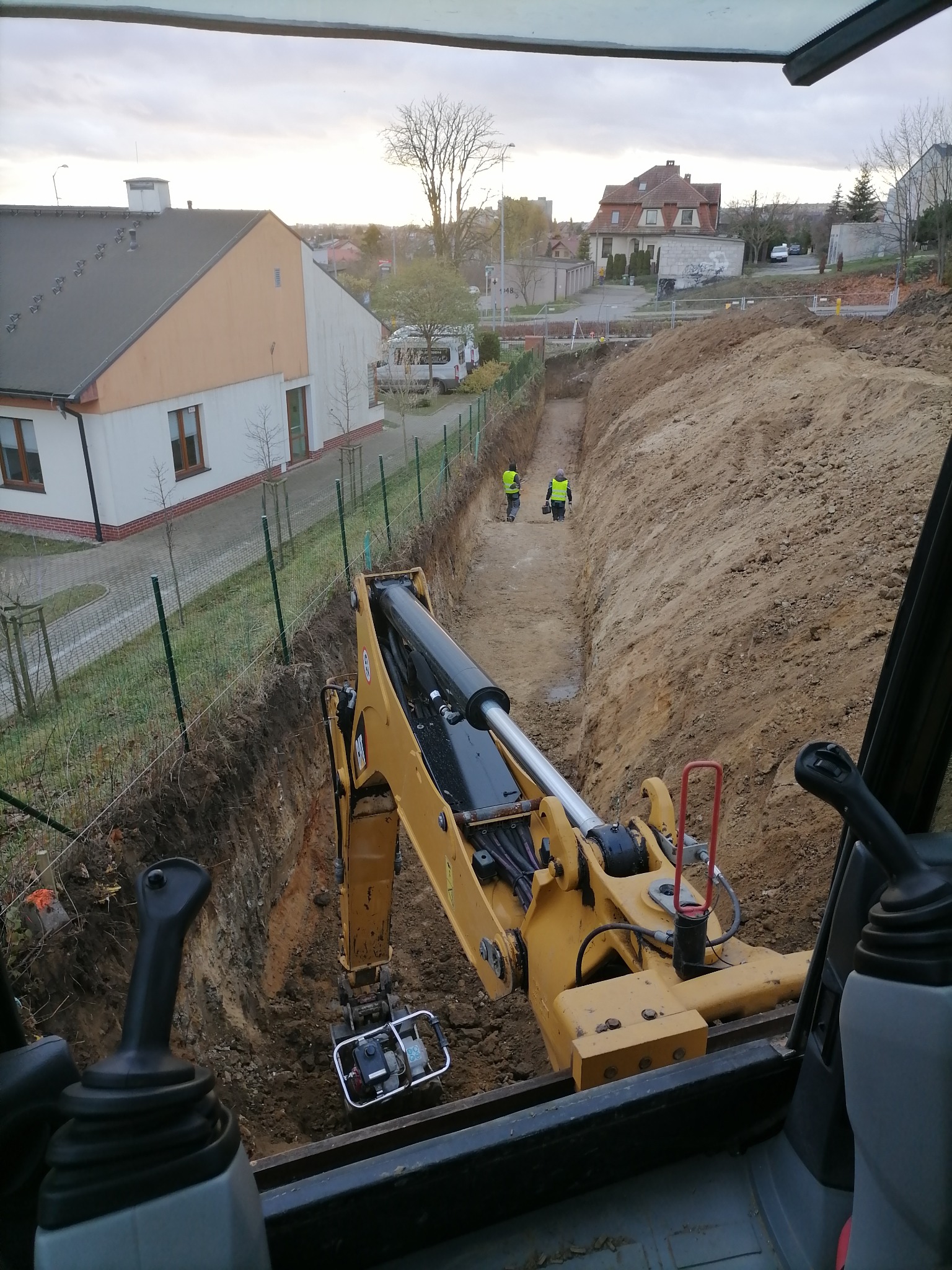 Żółta koparka z ubijarką w wykopie, widok z kabiny operatora, dwóch pracowników w kamizelkach odblaskowych w oddali, domy w tle.