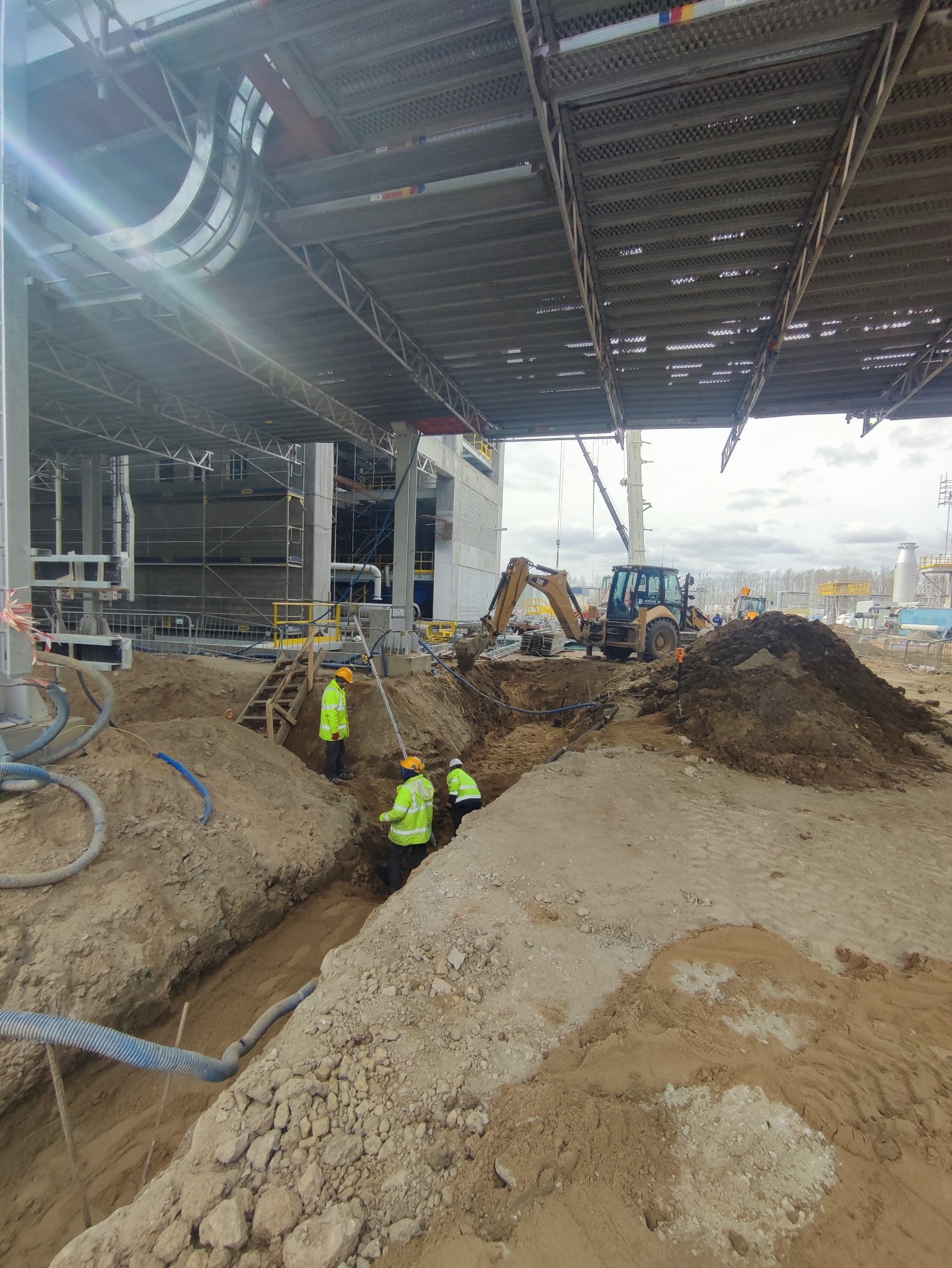 Pracownicy w jaskrawozielonych kamizelkach kopią wąski rów na placu budowy, w tle koparka i elementy konstrukcyjne budynku.