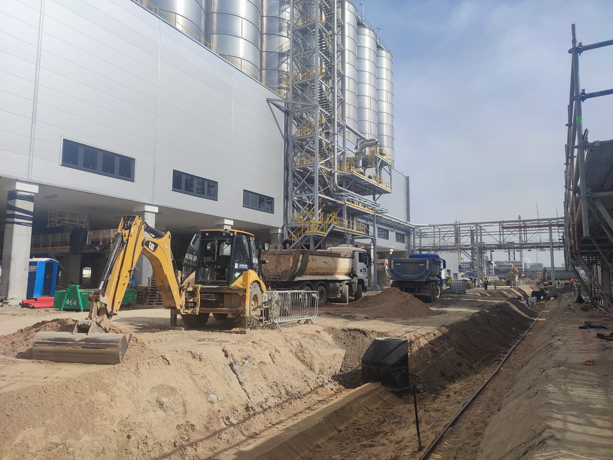 Żółta koparka CAT i wywrotki na placu budowy z wykopem, w tle silosy i konstrukcja stalowa, widoczne elementy infrastruktury i pracownicy.