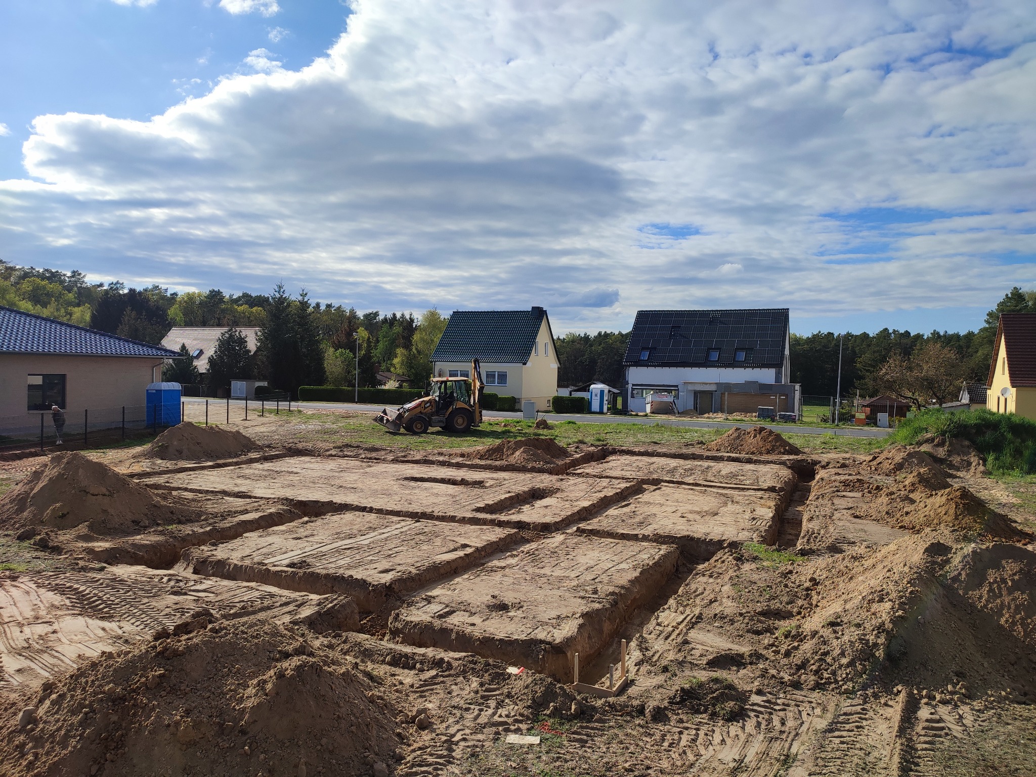 Przygotowany wykop pod fundamenty budynku mieszkalnego, widoczne ślady opon koparki i pryzmy ziemi, w tle zabudowa jednorodzinna i błękitne niebo z chmurami.