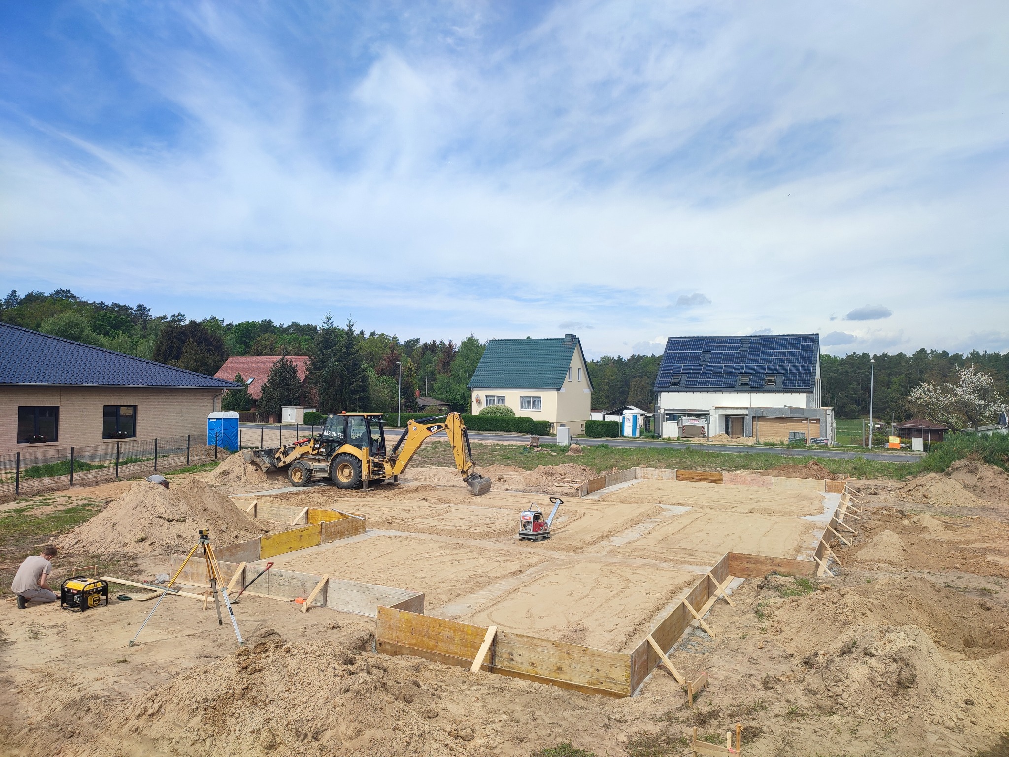 Przygotowanie terenu pod fundamenty domu jednorodzinnego: wykopane i utwardzone podłoże piaskowe w drewnianym szalunku, widoczny sprzęt budowlany - koparko-ładowarka i zagęszczarka, niwelator...