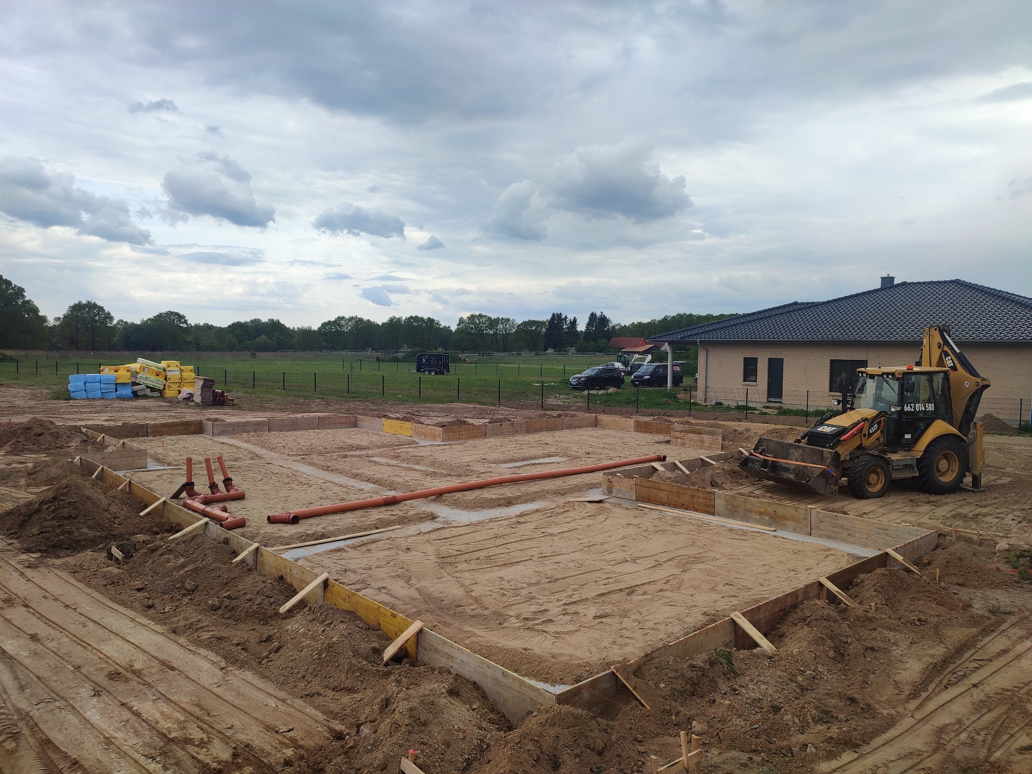 Przygotowanie fundamentów pod budynek jednorodzinny: wykop z drewnianym szalunkiem, ułożone rury kanalizacyjne, żółta koparko-ładowarka CAT i materiały budowlane w tle, pochmurne niebo.