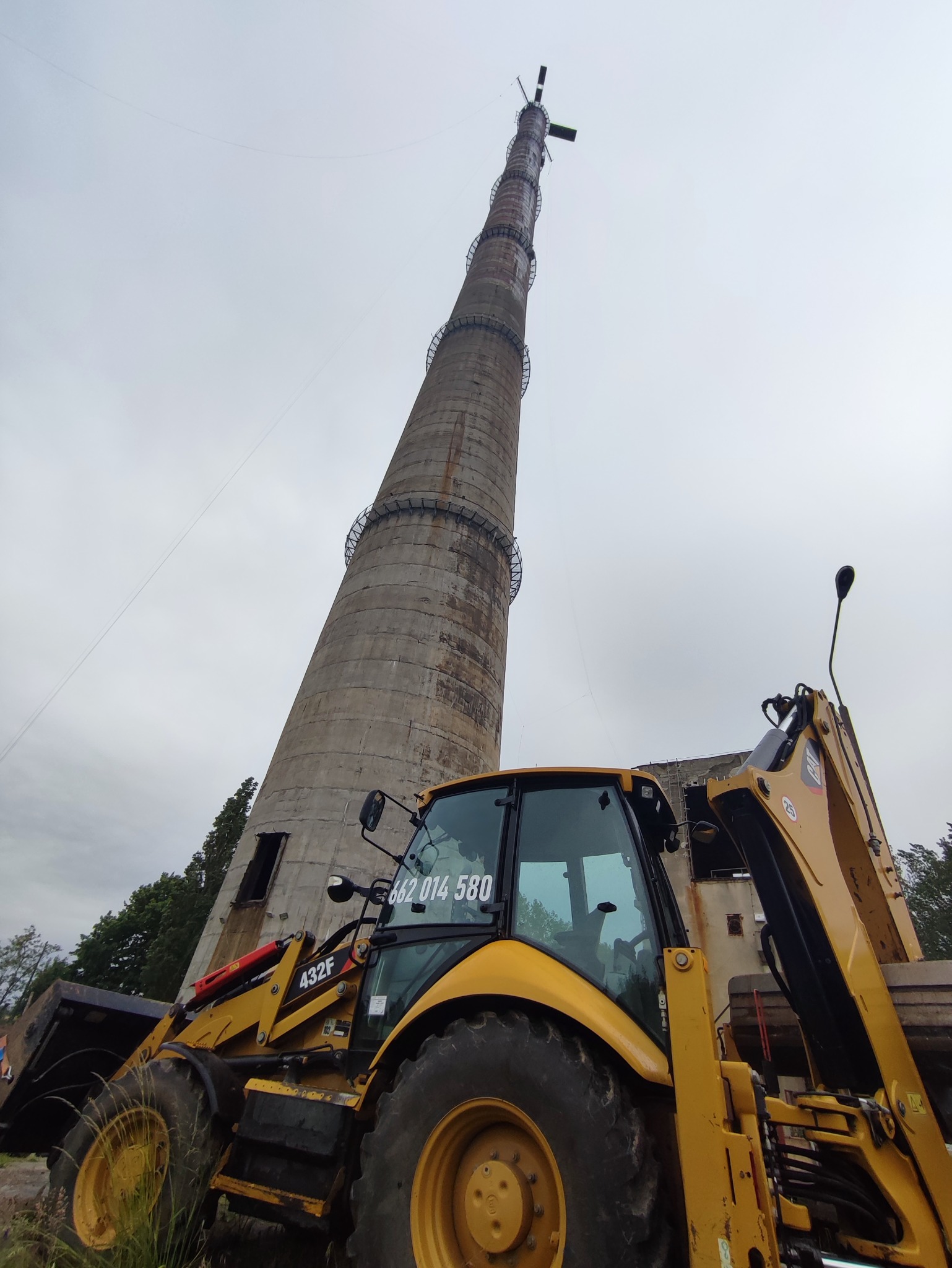 Żółta ładowarka teleskopowa Caterpillar 432F na tle wysokiego, betonowego komina z metalowymi schodami, pochmurne niebo.