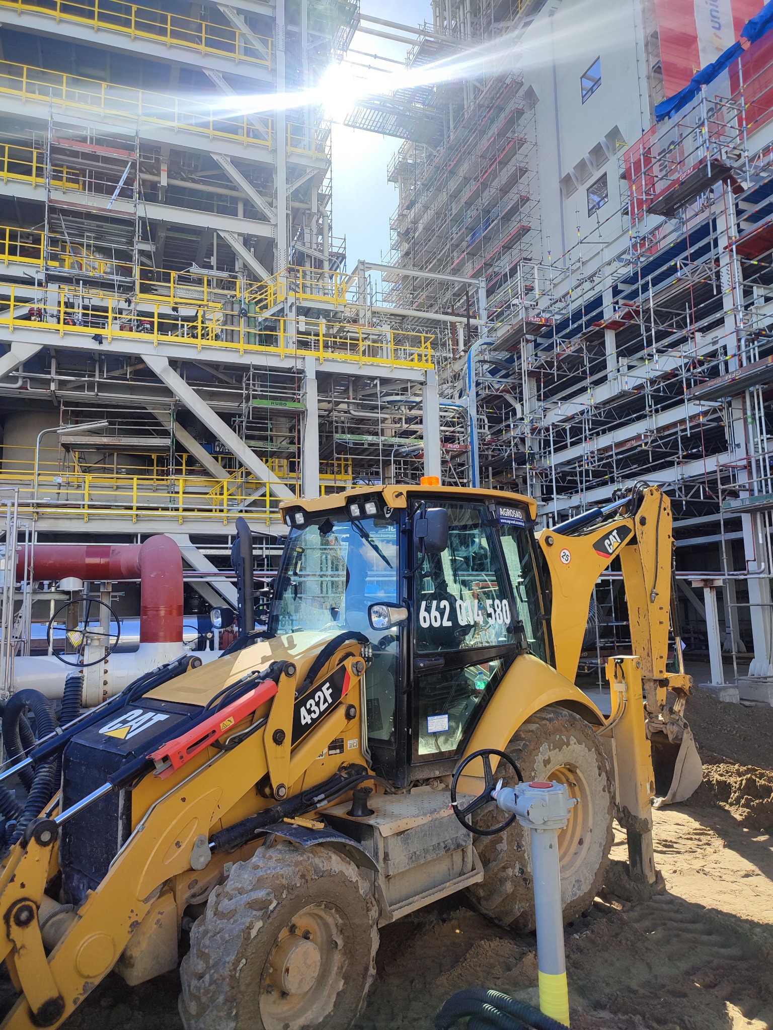 Żółta koparko-ładowarka CAT 432F na placu budowy, z widocznymi rusztowaniami i elementami konstrukcyjnymi w tle, ziemia wokół maszyny, przewody i zawór w pobliżu koła.