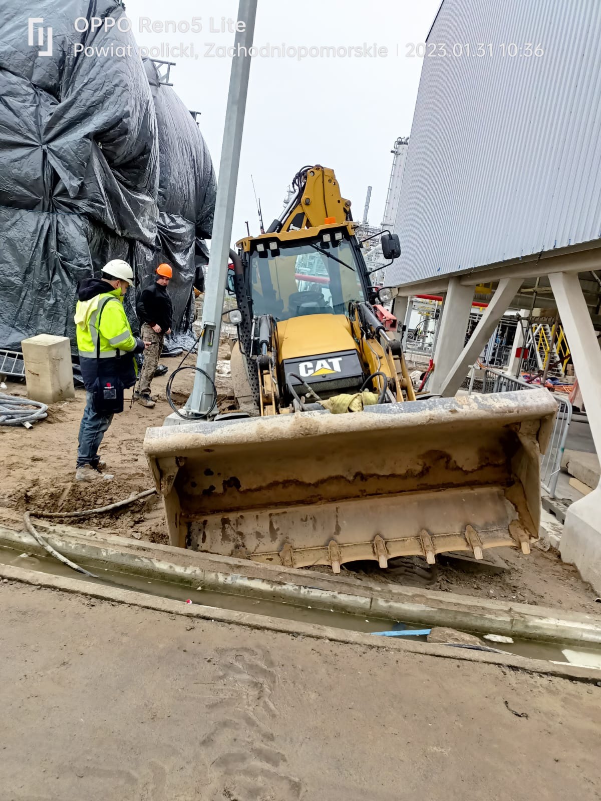 Żółta koparko-ładowarka CAT na placu budowy, z widocznymi pracownikami w odblaskowych kamizelkach, na tle konstrukcji stalowej i czarnej folii ochronnej.