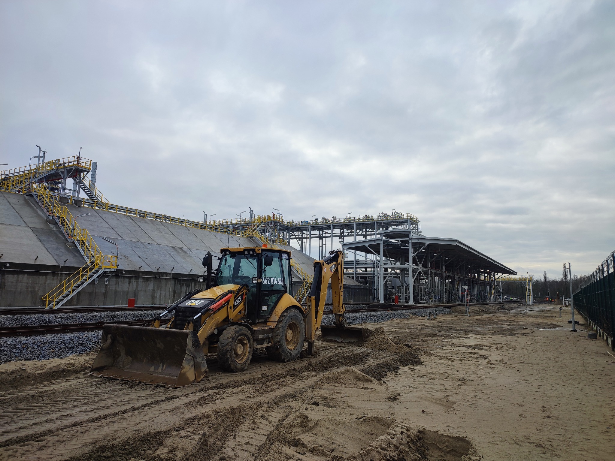 Żółta koparko-ładowarka CAT 432F na placu budowy, widoczne tory kolejowe i instalacje przemysłowe w tle pod pochmurnym niebem.