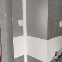 Wykończona ściana w łazience z szarą, strukturalną gładzią imitującą kamień, przecięta poziomym, białym pasem, z widocznym fragmentem chromowanego uchwytu i włącznikiem światła.