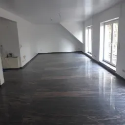 Przestronne, jasne wnętrze z nową, ciemną, polerowaną podłogą imitującą marmur, świeżo pomalowane białe ściany i sufity, okna z widokiem na zewnątrz.