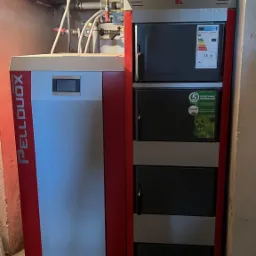 Nowoczesny, dwuczęściowy kocioł grzewczy z podajnikiem, marki Kolton Pellduox, umieszczony na betonowych bloczkach w piwnicy, z widocznymi rurami instalacji grzewczej pod sufitem.