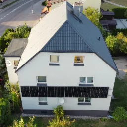 Widok z góry na dom z panelami słonecznymi na elewacji, szarym dachem i anteną satelitarną. W tle widoczna ulica i inne budynki.