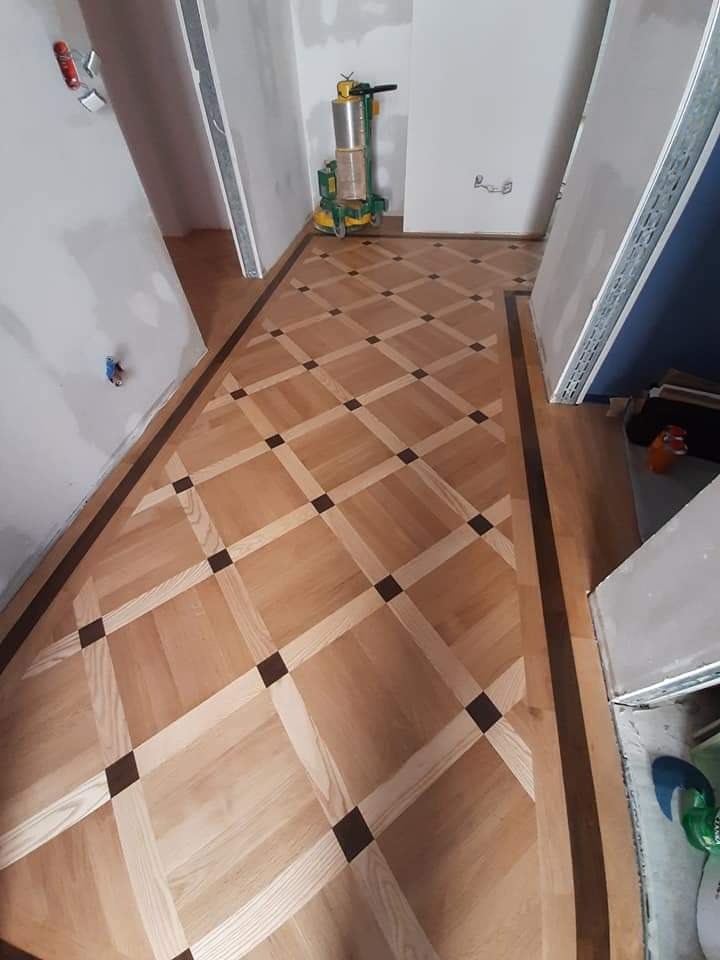 Wzór parkietu w romby z ciemnymi akcentami, widoczna maszyna do cyklinowania parkietu w tle, ściany w trakcie remontu.