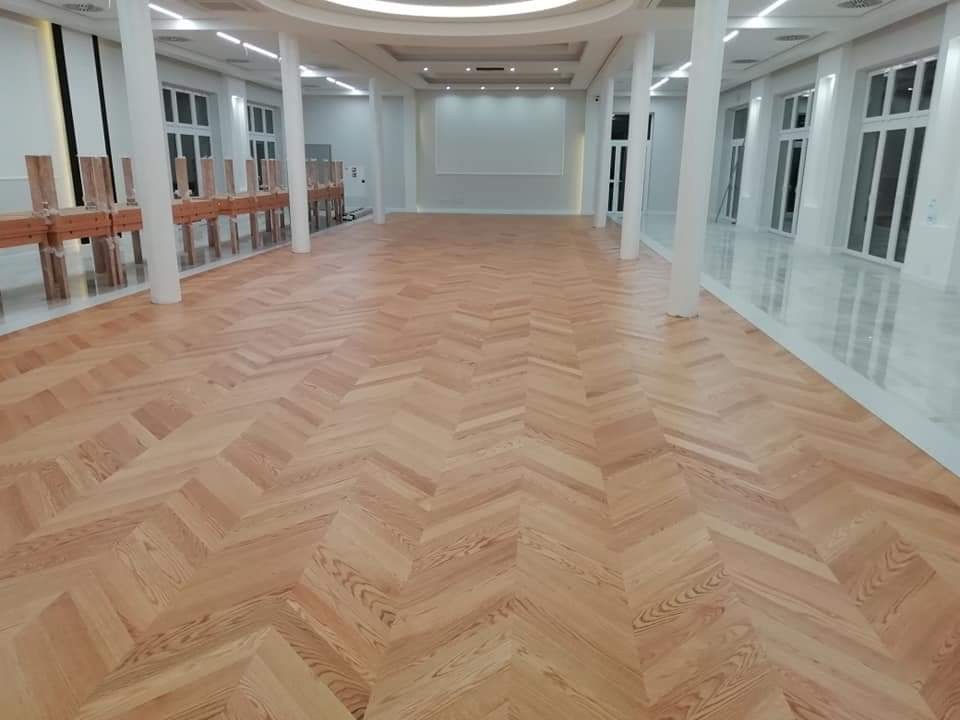 Jasny parkiet ułożony we wzór jodełki w przestronnym, białym wnętrzu z kolumnami i dużymi oknami.