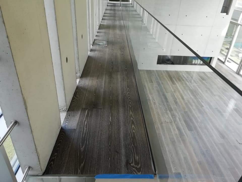 Nowoczesne wnętrze z dwiema strefami podłogowymi: ciemne deski w korytarzu i jasne w salonie, oddzielone szklaną balustradą.