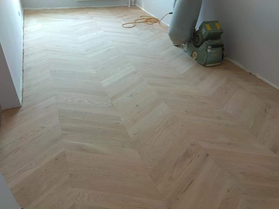 Parkiet ułożony we wzór jodełki francuskiej, świeżo po cyklinowaniu, z widoczną maszyną do cyklinowania przy ścianie.