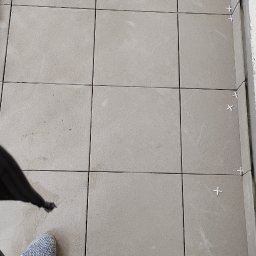 Tomasz Czaja - Beżowe płytki podłogowe w trakcie układania, widoczne krzyżyki dystansowe, ujęcie z góry z fragmentem butów i nogawki spodni glazurnika.