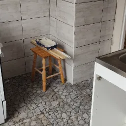 Ściana w kuchni wyłożona szarymi płytkami imitującymi drewno. Na drewnianym taborecie ustawiona kuweta malarska i listwa pomiarowa. Na podłodze płytki imitujące kamień. Widoczny fragment szafki...