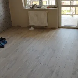 Pokój w trakcie remontu: ułożone panele podłogowe w odcieniach szarości, widoczne materiały budowlane i torby z ubraniami przy ścianie z tapetą, grzejnik i okno w tle, brak listew przypodłogowych.