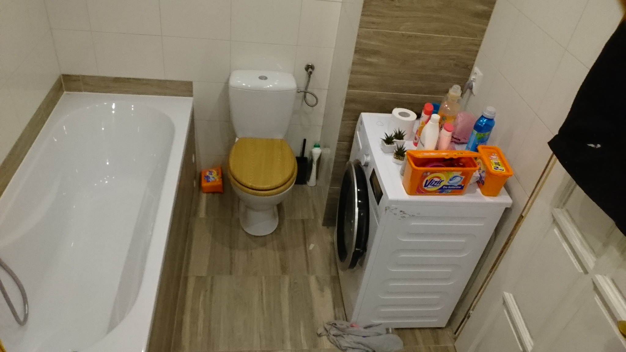 Wnętrze łazienki z białą wanną, toaletą z drewnianą deską, pralką i różnymi środkami czystości na niej