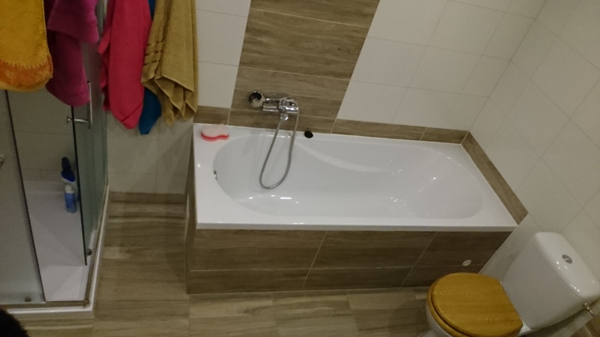 Wykończona łazienka z białą wanną obudowaną płytkami imitującymi drewno, prysznicem ze szklanymi drzwiami i toaletą z drewnianą deską, widoczna armatura i akcesoria łazienkowe.