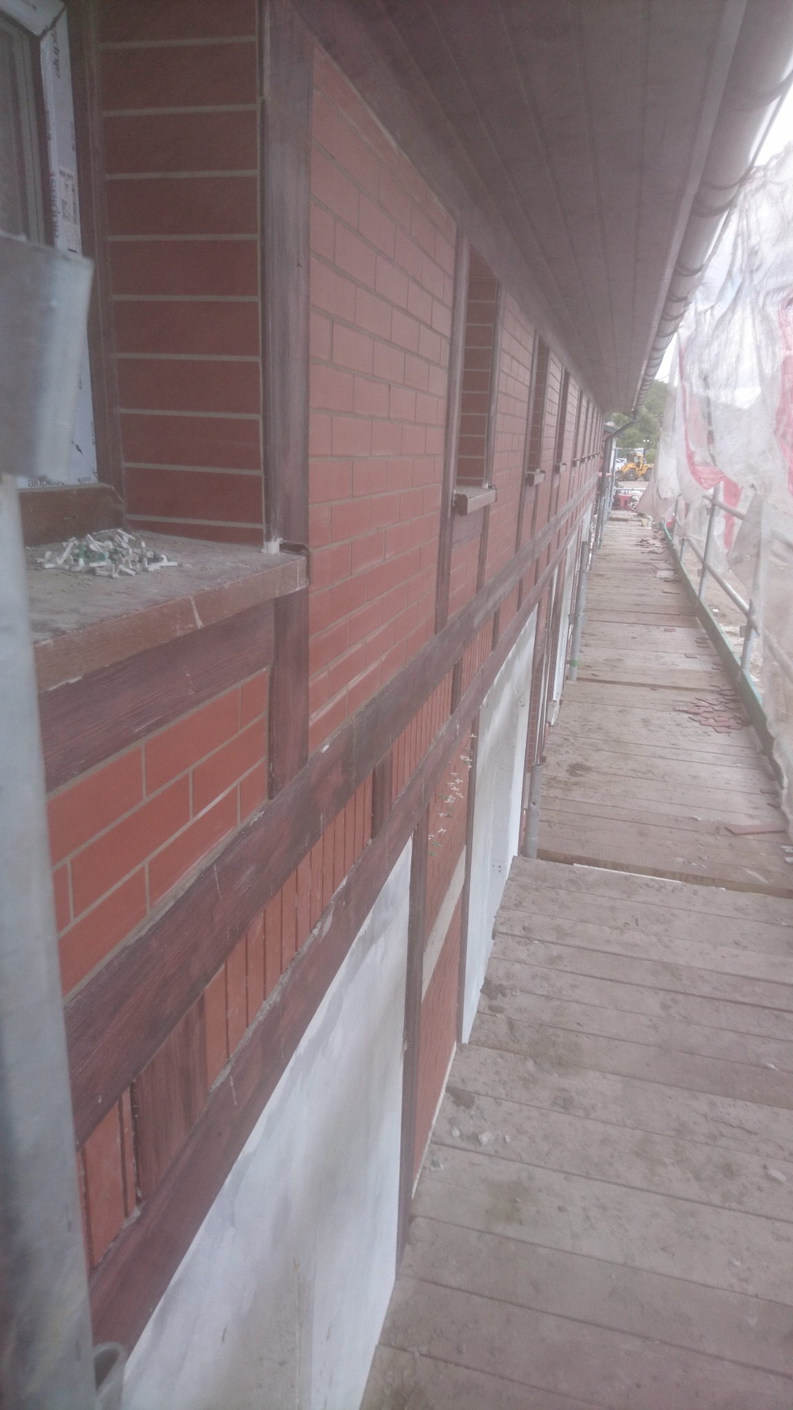 Ściana budynku w trakcie prac murarskich, widoczne cegły, drewniane elementy konstrukcyjne, rusztowanie i materiały budowlane na parapecie okiennym.