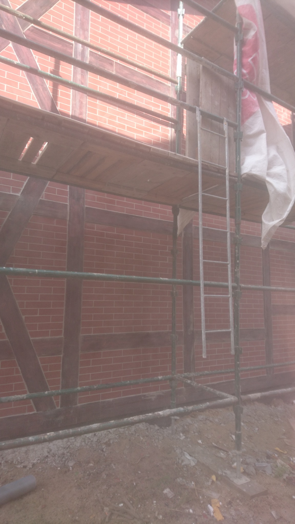 Częściowe ujęcie rusztowania przed budynkiem z czerwonej cegły z drewnianą konstrukcją szkieletową, widoczna drabina i fragment plandeki ochronnej.