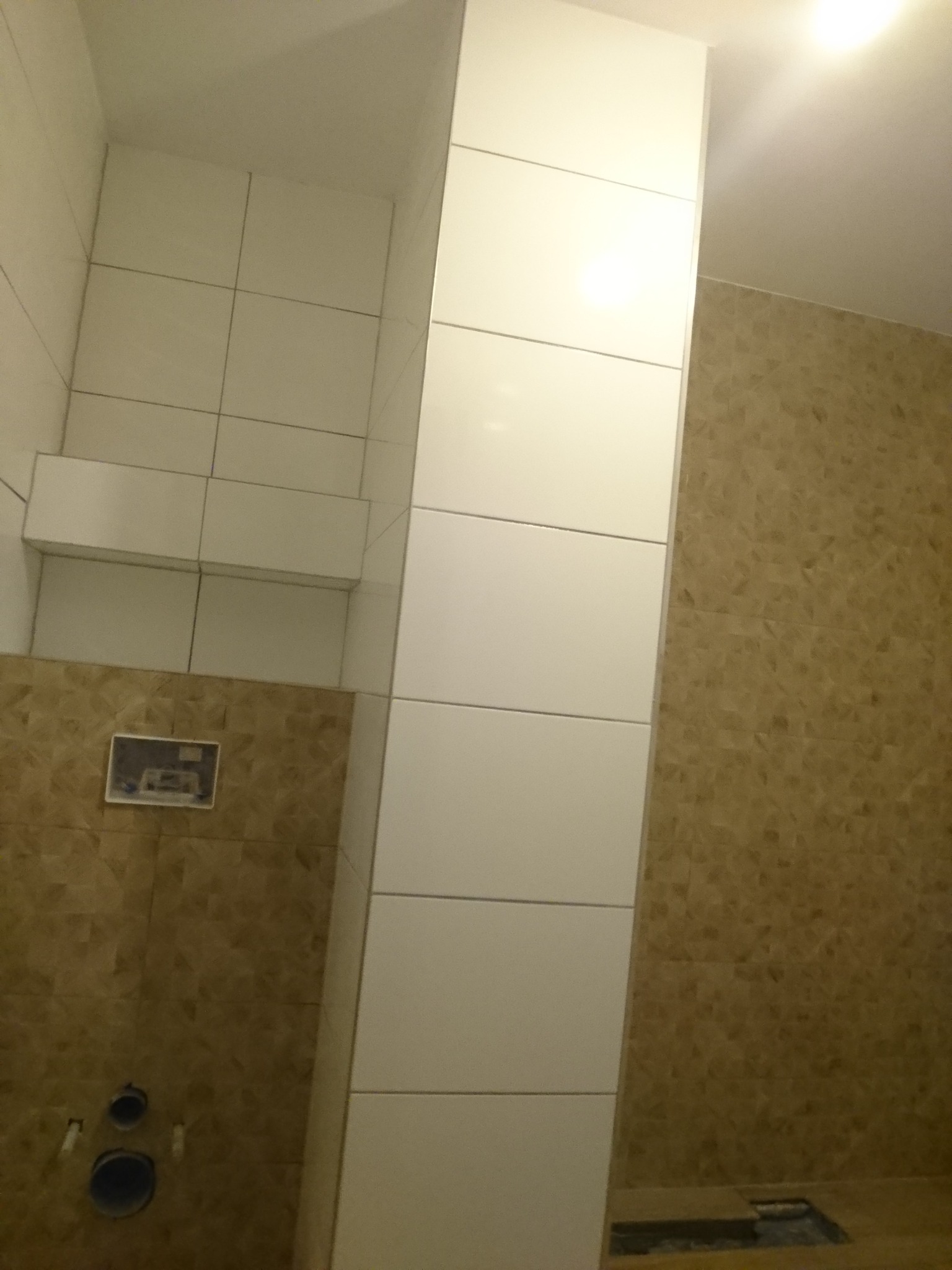 Wykończona łazienka z brązowymi i białymi płytkami, widoczne otwory na rury i zabudowa stelaża WC.