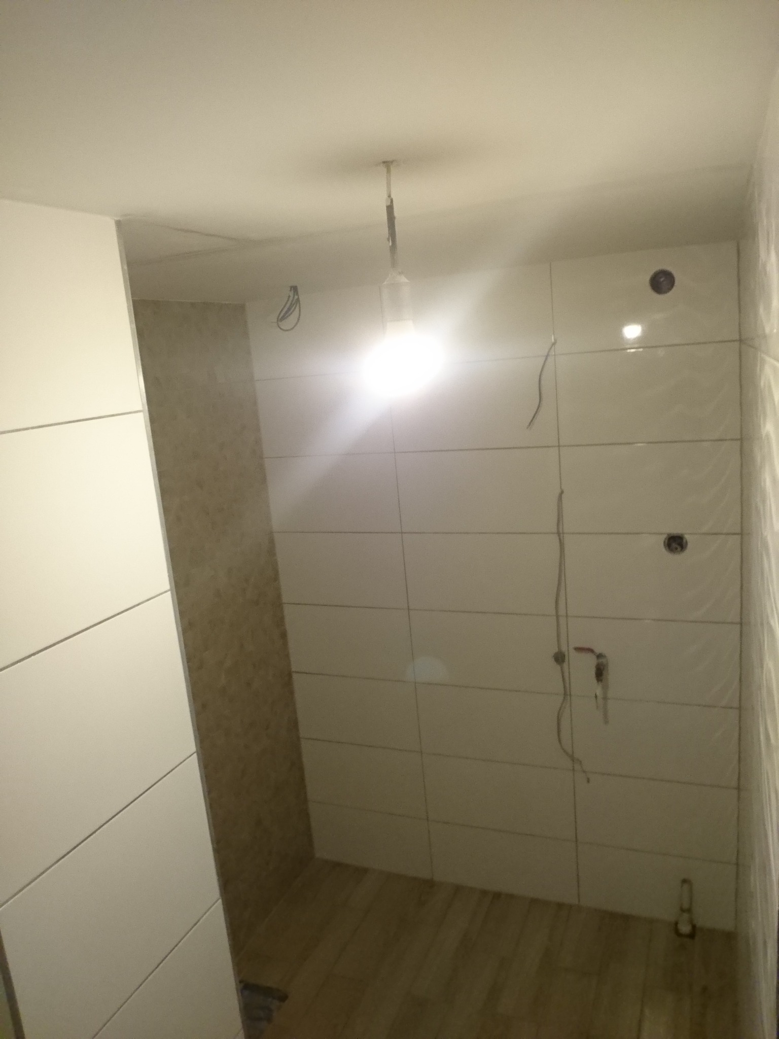 Wnętrze łazienki w trakcie prac wykończeniowych; widoczne białe, błyszczące płytki ułożone na ścianach, wystające przewody elektryczne, surowa żarówka na kablu oraz fragment podłogi...