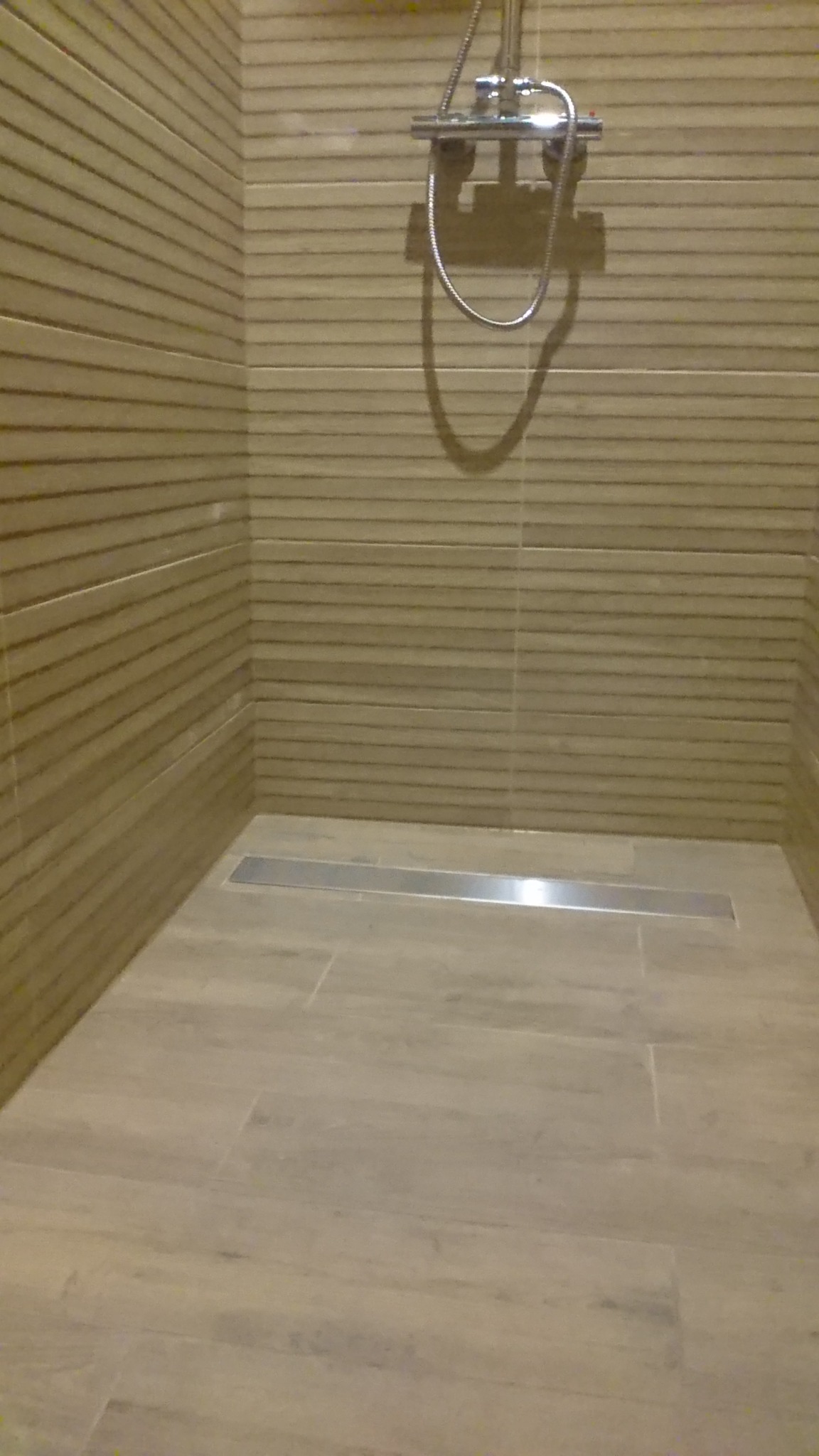 Nowoczesna kabina prysznicowa z odpływem liniowym, wykończona płytkami imitującymi drewno, z chromowaną armaturą.