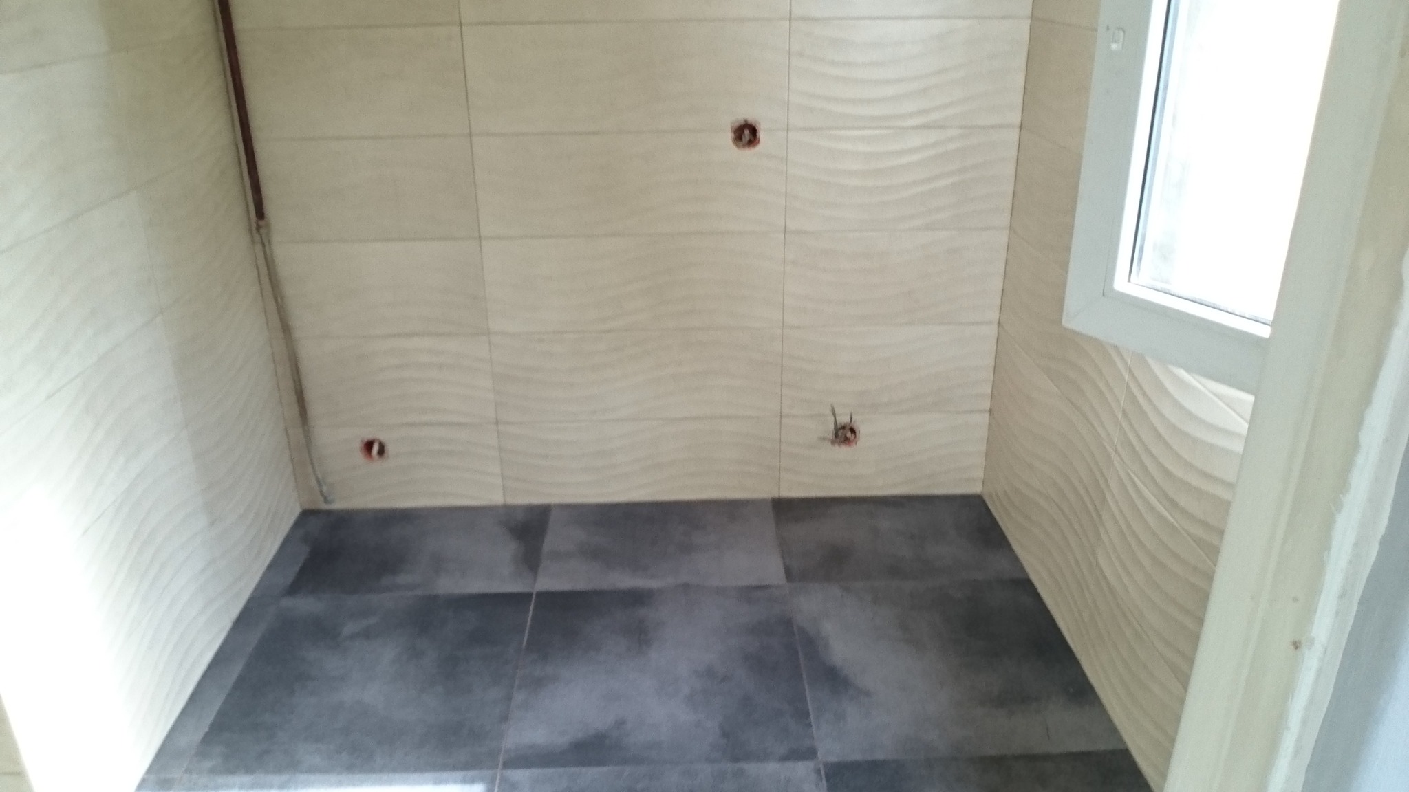 Świeżo wykafelkowana łazienka z beżowymi płytkami ściennymi o falistej strukturze i ciemnoszarą podłogą z płytek. Widoczne przygotowane punkty elektryczne.