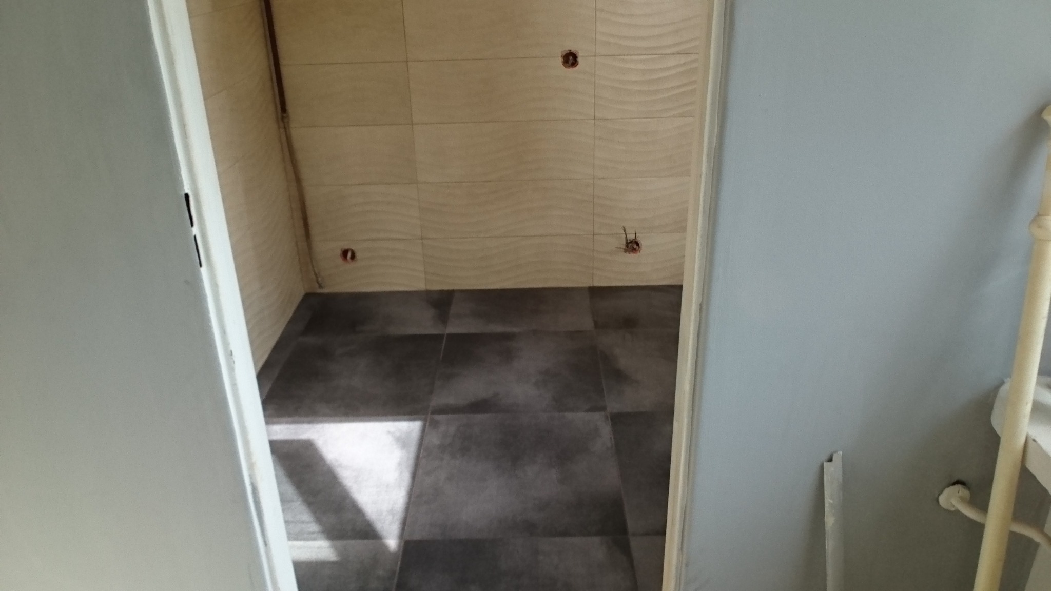 Łazienka w trakcie wykańczania: położone ciemne płytki podłogowe, jasne płytki ścienne z delikatnym wzorem, widoczne puszki elektryczne bez osprzętu.