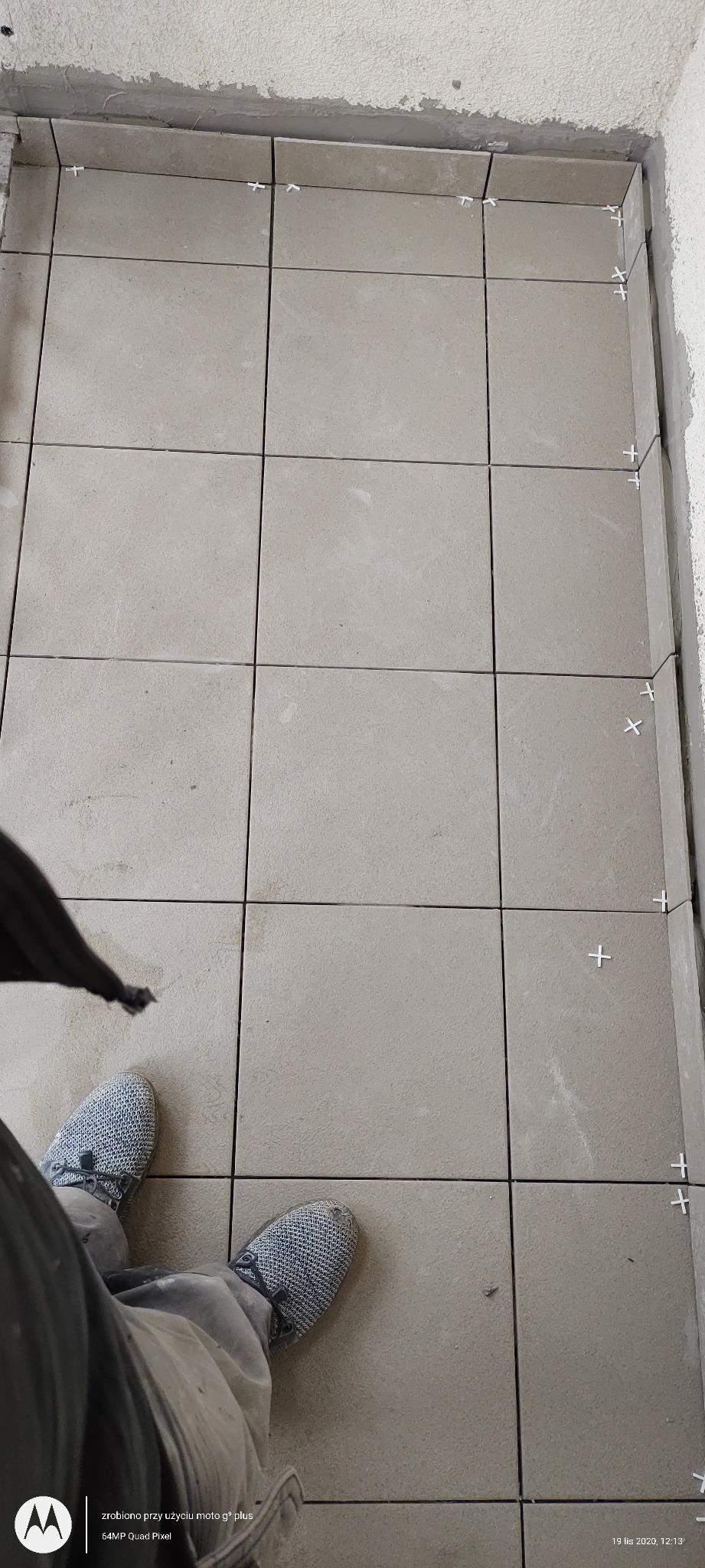 Beżowe płytki podłogowe w trakcie układania, widoczne krzyżyki dystansowe, ujęcie z góry z fragmentem butów i nogawki spodni glazurnika.