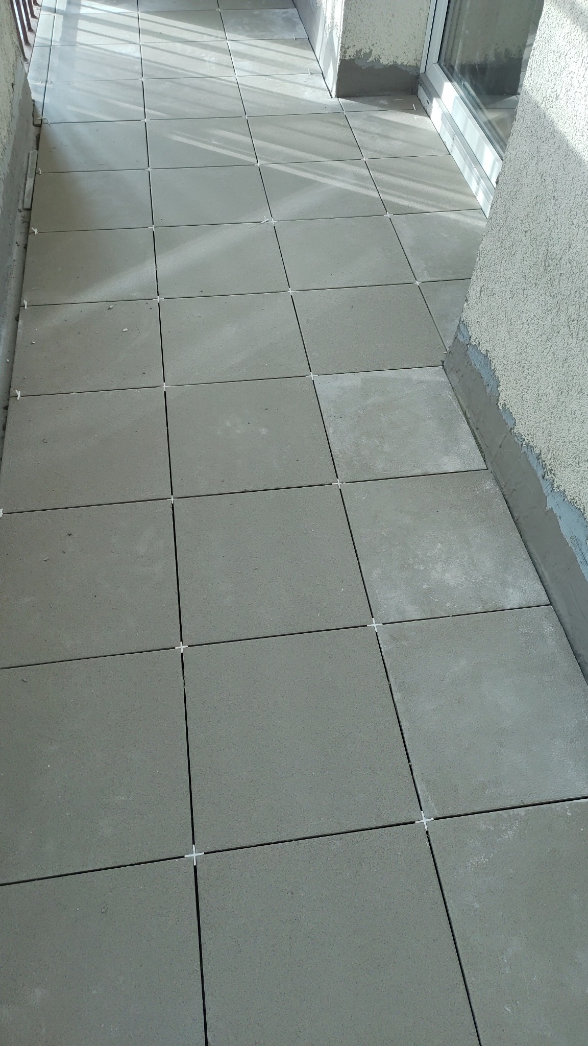 Świeżo ułożone, szare płytki na balkonie, z krzyżykami dystansowymi w spoinach, widoczne surowe wykończenie ścian.