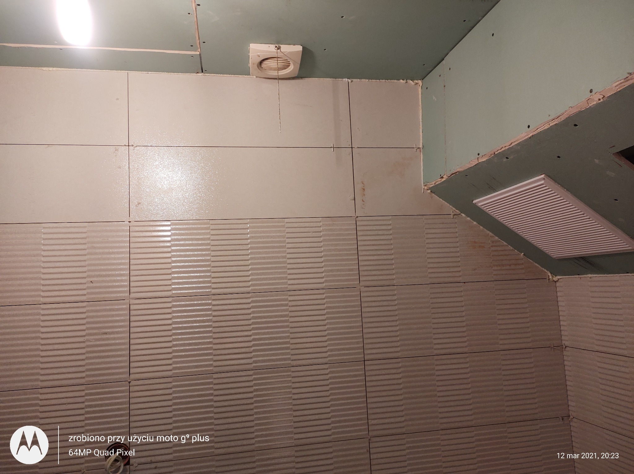 Ściana w trakcie układania płytek z poziomym wzorem, widoczne krzyżyki dystansowe, sufit z płyt gipsowo-kartonowych i kratka wentylacyjna.