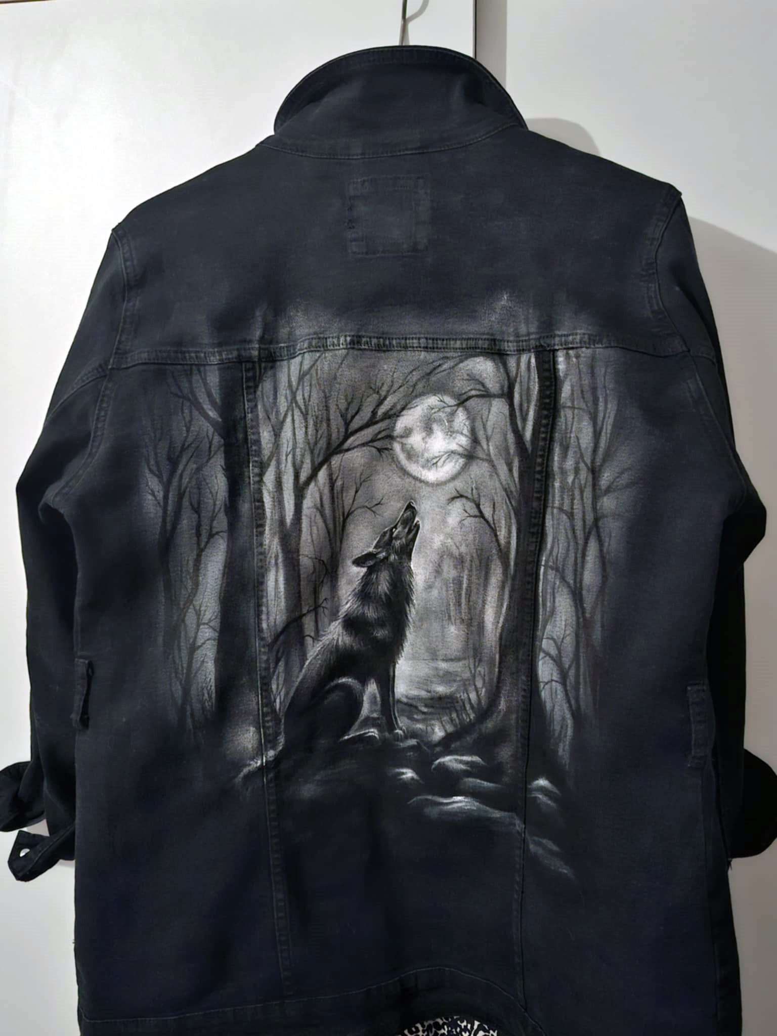 Czarna kurtka jeansowa z ręcznie malowanym motywem wilka wyjącego do księżyca w lesie, wykonana w czerni i bieli. Unikatowy projekt artystyczny na odzieży.