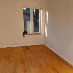 Deco8 - Puste, jasne pomieszczenie z drewnianą podłogą po cyklinowaniu, widoczne ślady po procesie, białe ściany, okno z widokiem na budynek gospodarczy.