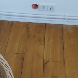 Deco8 - Niedokończone gniazdko elektryczne w ścianie nad drewnianą podłogą, widoczna puszka instalacyjna i dwa gniazdka obok, białe listwy przypodłogowe.