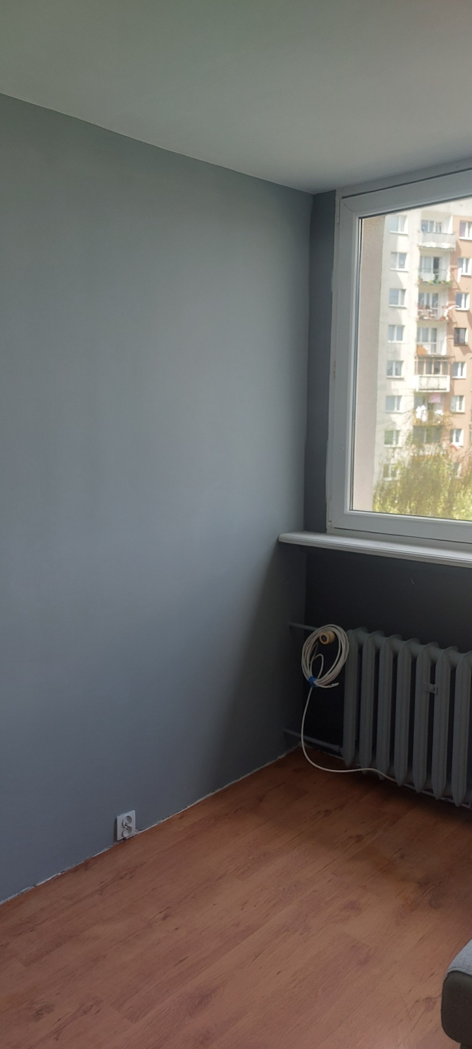 Pomalowane na szaro wnętrze pokoju z grzejnikiem i widokiem na blok przez okno, brązowe panele na podłodze.