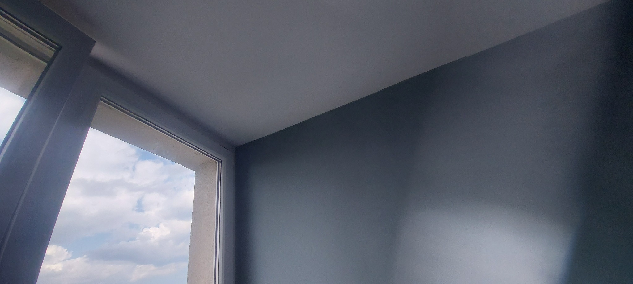 Pomalowany na szaro róg pokoju z oknem, widok na niebo z chmurami.
