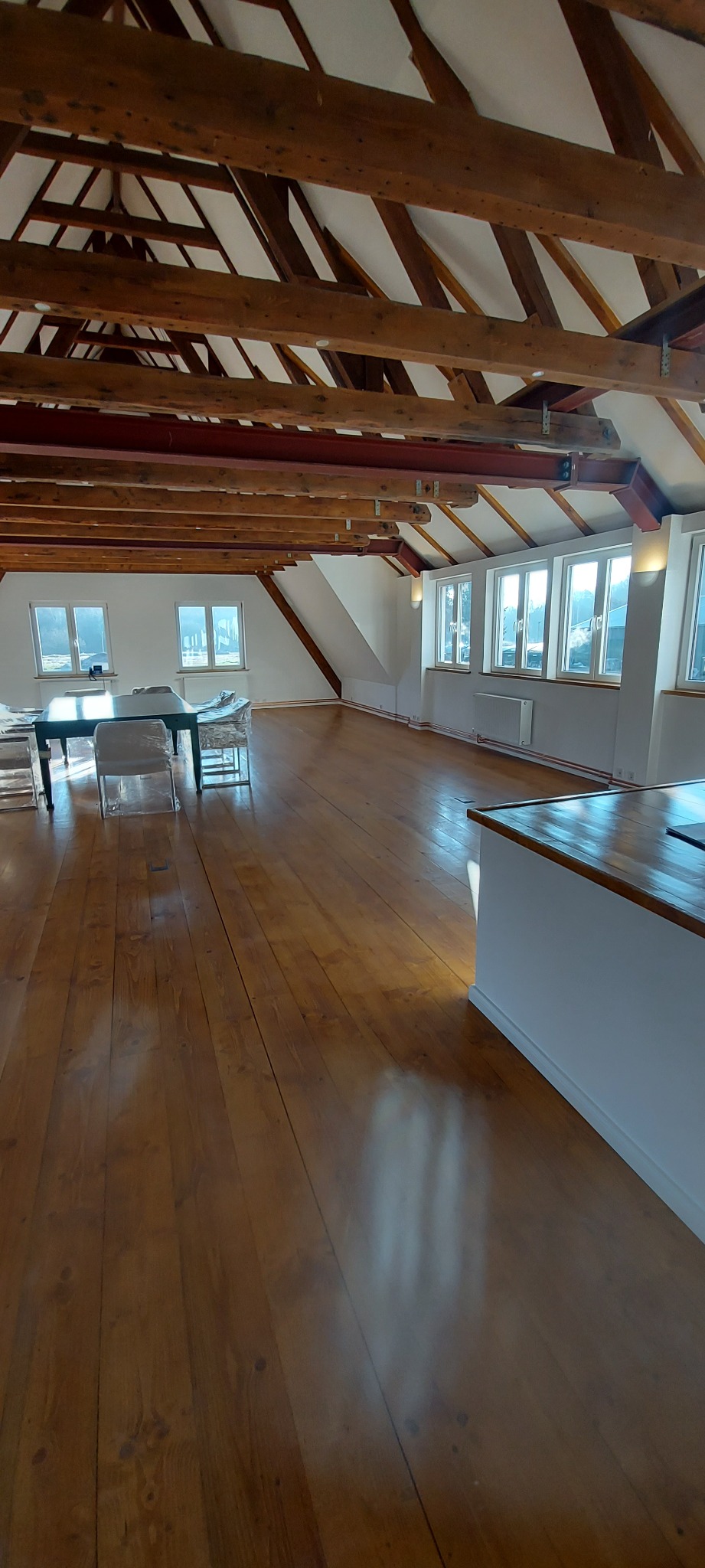 Przestronne poddasze z drewnianą podłogą i odsłoniętą konstrukcją więźby dachowej, okna w białych ramach wpuszczają naturalne światło, meble zabezpieczone folią ochronną.