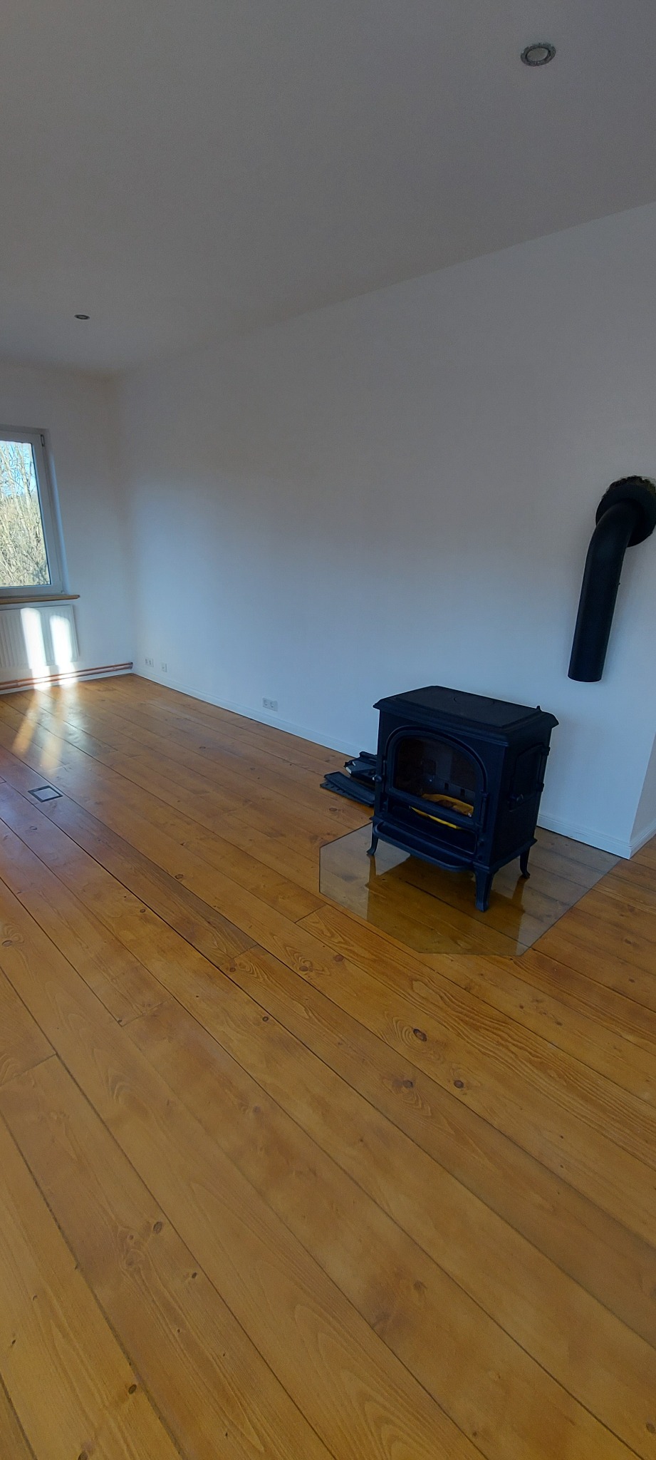 Wnętrze pokoju z drewnianą podłogą po cyklinowaniu, widoczny piec wolnostojący z rurą odprowadzającą dym, białe ściany i okno z widokiem na drzewa.