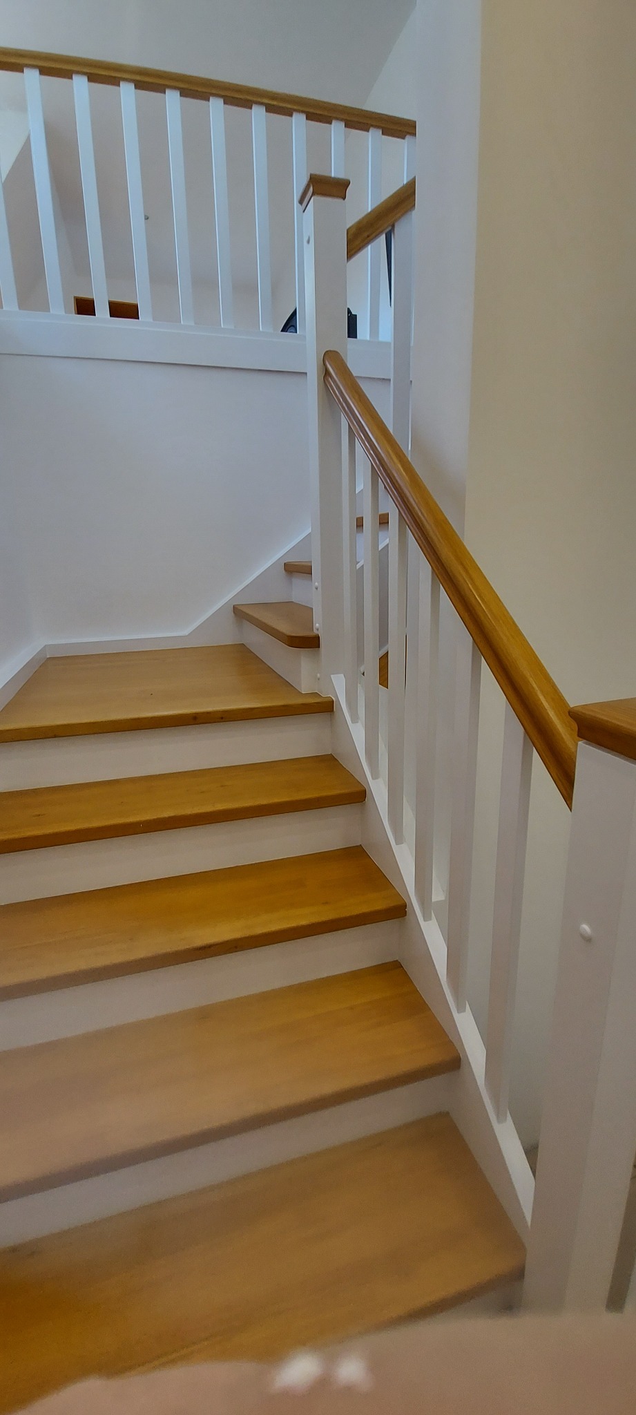 Jasne, drewniane schody z białymi balustradami i poręczą w odcieniu naturalnego drewna, widok z dołu, ukazujący detale wykończenia i gładką powierzchnię drewna.