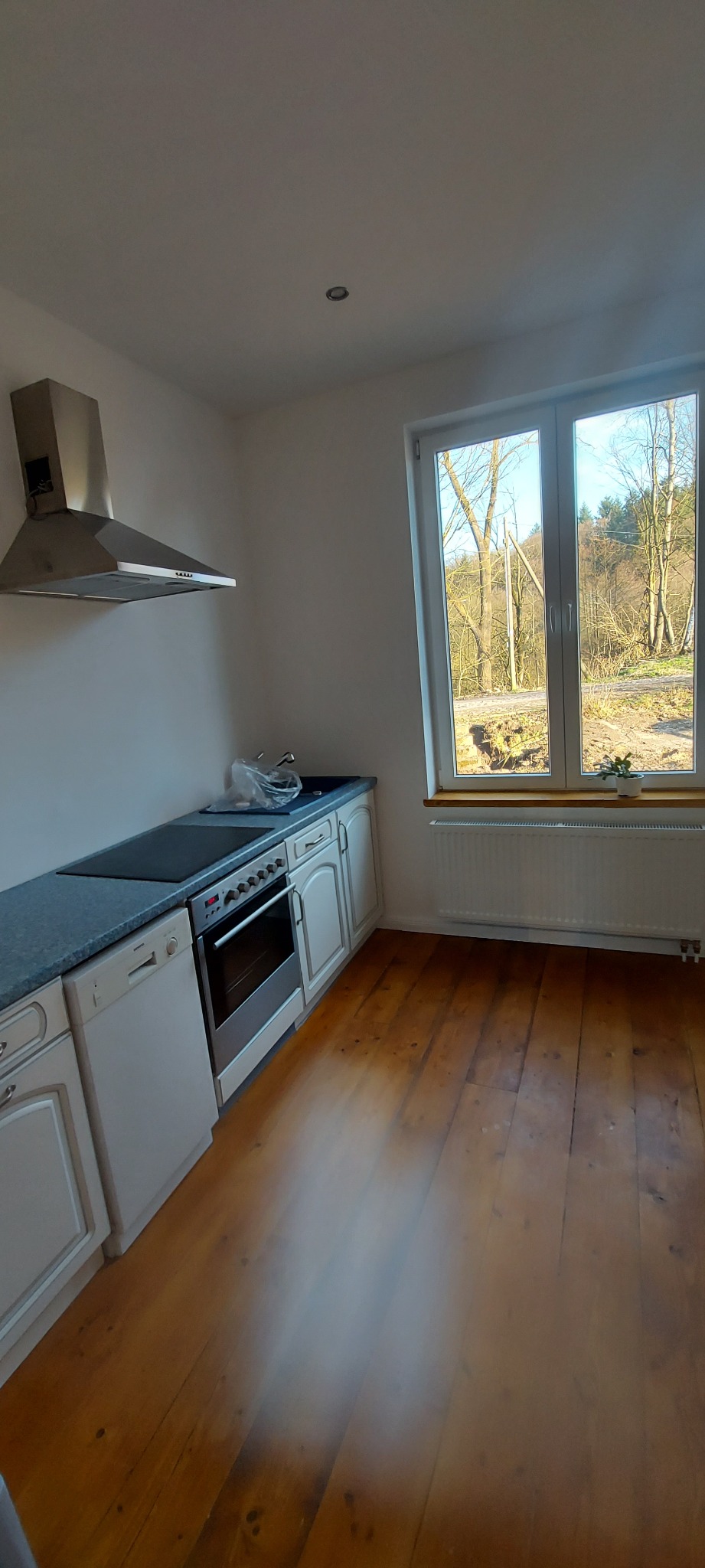 Jasna kuchnia z drewnianą podłogą, białymi szafkami, piekarnikiem i oknem z widokiem na drzewa.