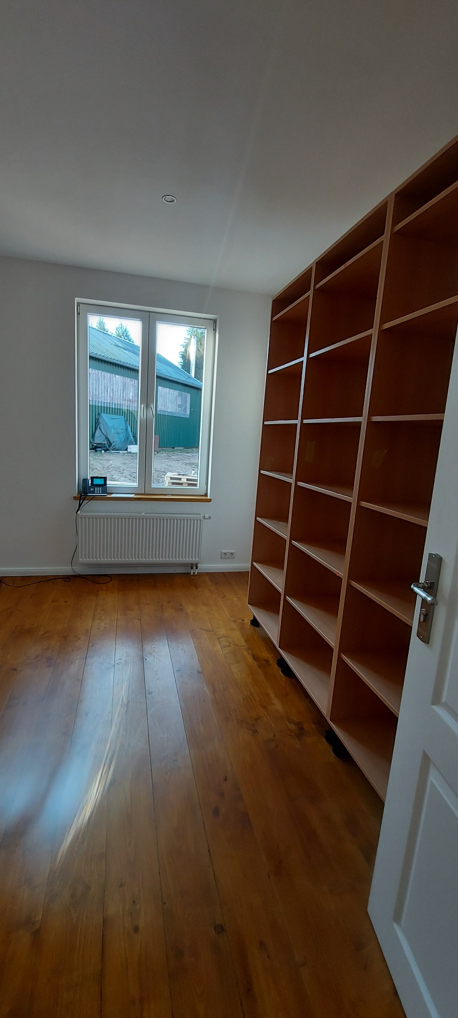 Pusty pokój z drewnianą podłogą, białymi ścianami i wysokim, drewnianym regałem na kółkach, stojącym przy ścianie. Widok na zewnątrz przez okno z grzejnikiem.