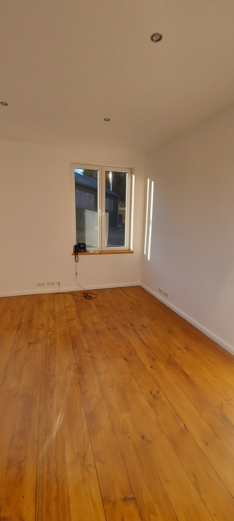 Puste, jasne pomieszczenie z drewnianą podłogą po cyklinowaniu, widoczne ślady po procesie, białe ściany, okno z widokiem na budynek gospodarczy.