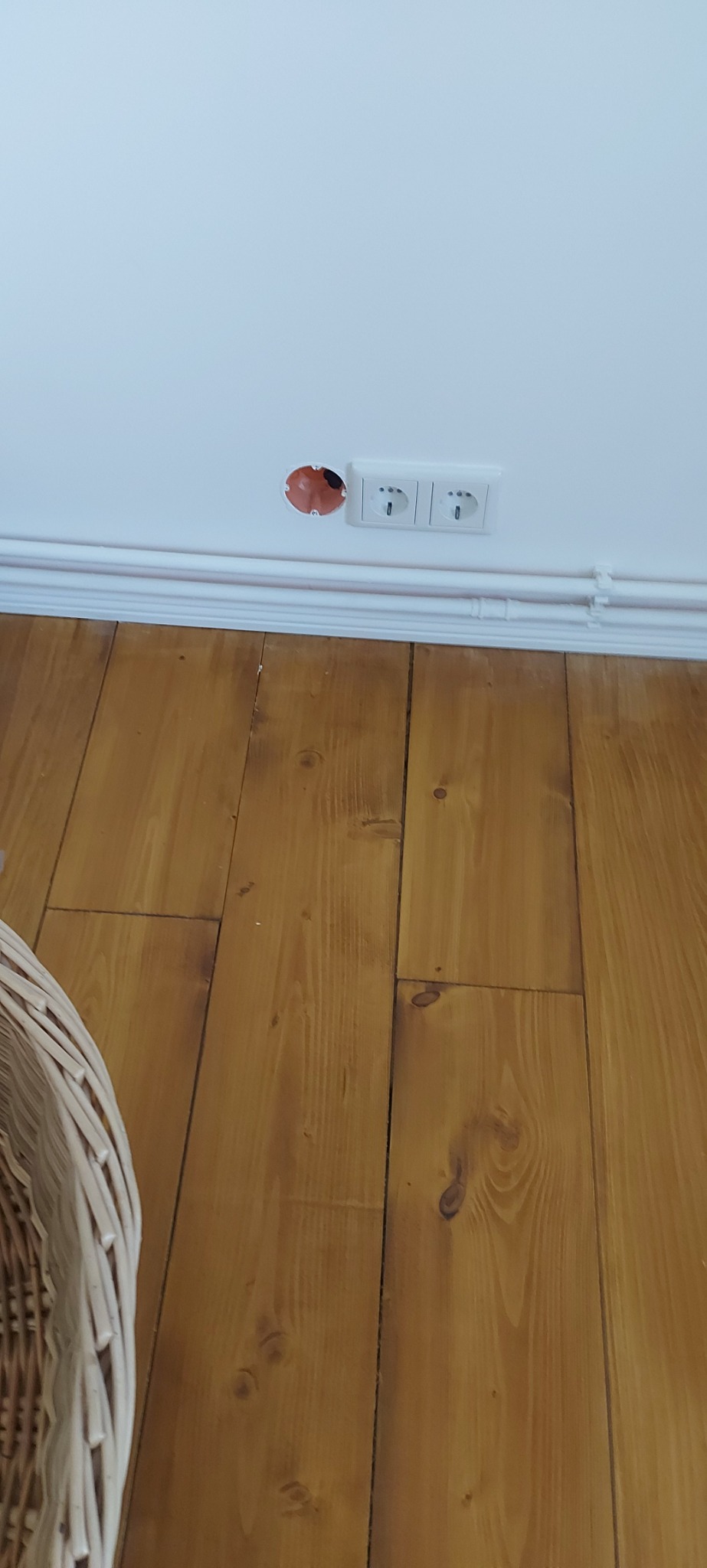 Niedokończone gniazdko elektryczne w ścianie nad drewnianą podłogą, widoczna puszka instalacyjna i dwa gniazdka obok, białe listwy przypodłogowe.