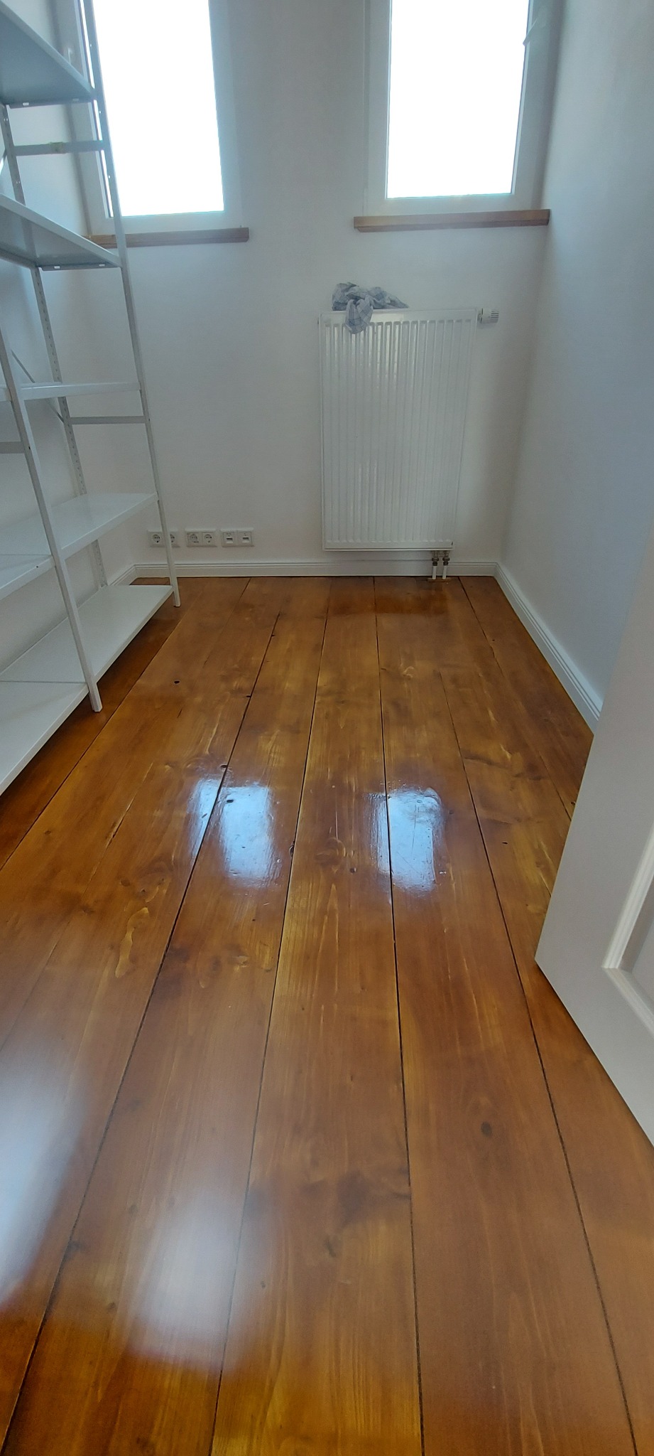 Wypolerowana, lakierowana podłoga z drewnianych desek w jasnym pomieszczeniu z białymi ścianami, widoczny biały grzejnik i biały regał.