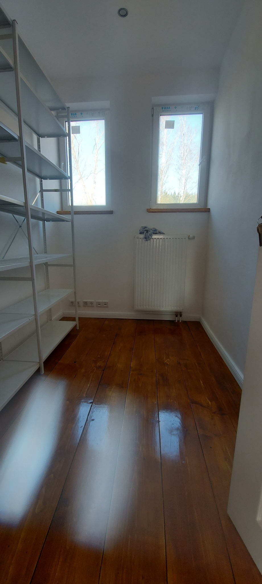 Pokój po cyklinowaniu z widoczną odnowioną, błyszczącą podłogą z desek, białymi ścianami, grzejnikiem pod oknem i metalowym regałem.