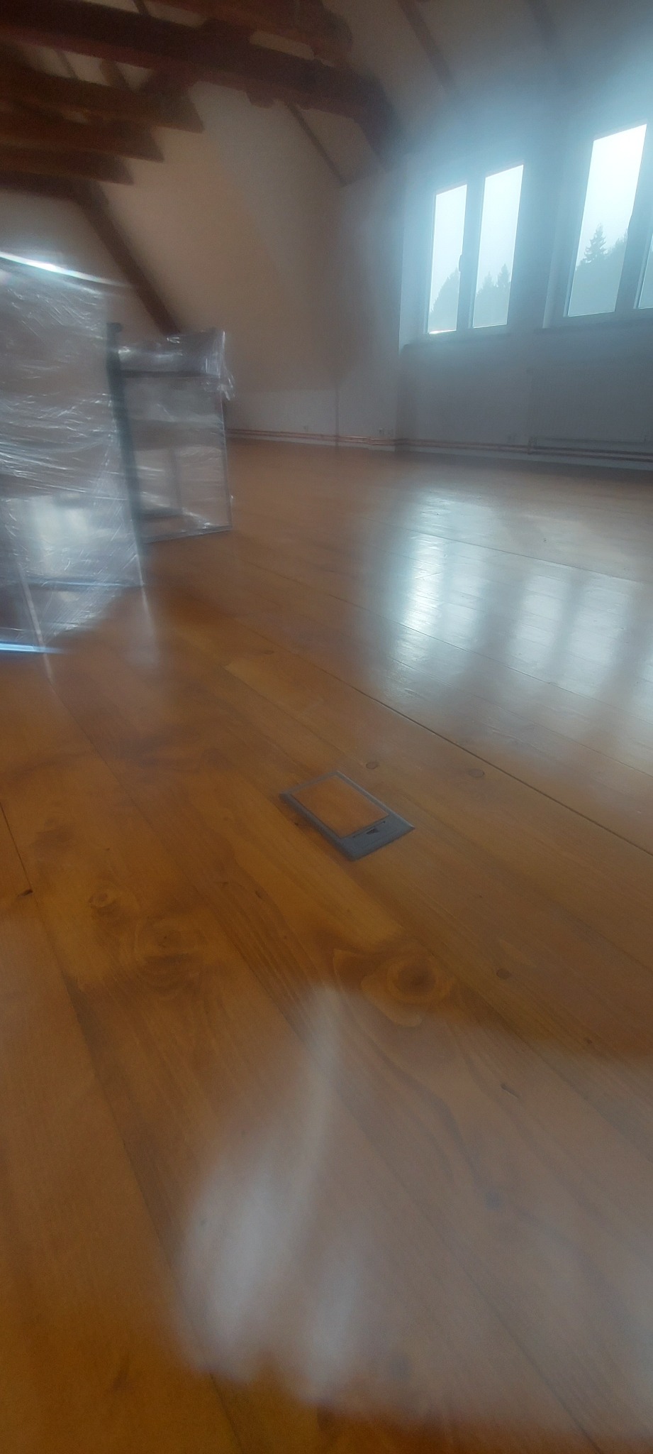 Błyszcząca, wycyklinowana podłoga z drewna w jasnym pomieszczeniu na poddaszu, widoczne okna i zabezpieczone meble folią.